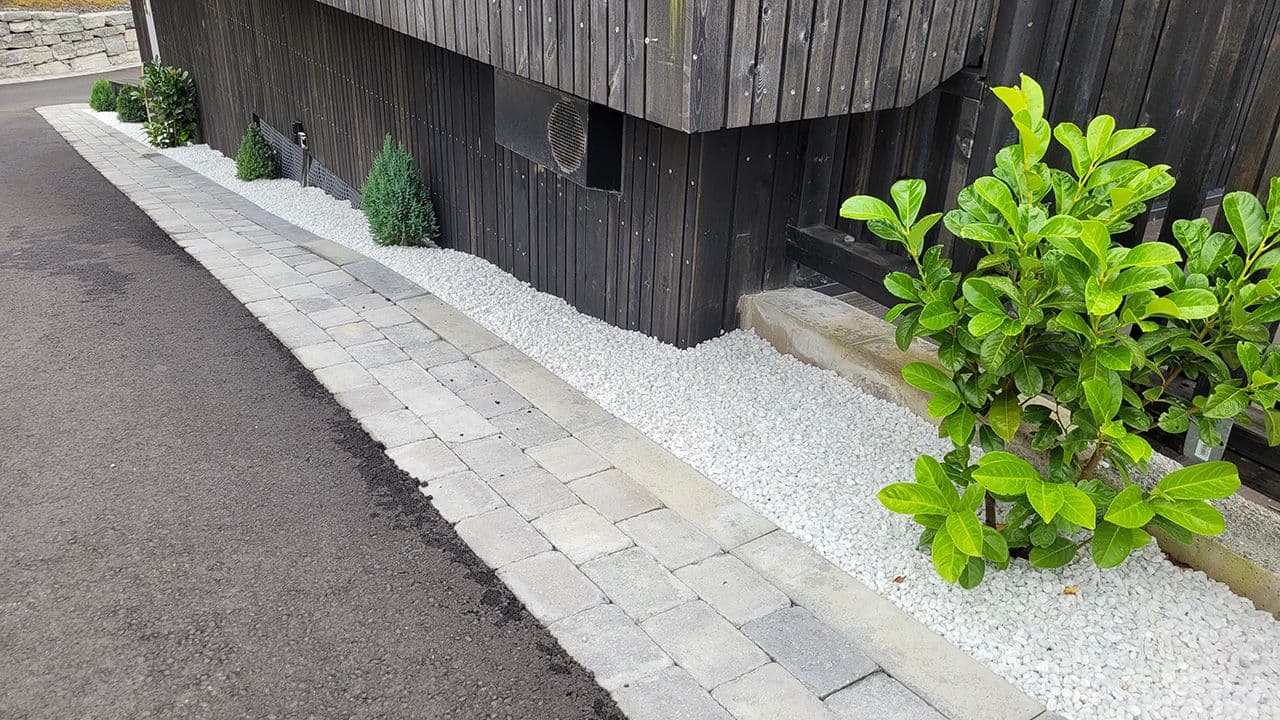 Dekorstein i blomsterbed langs svart husvegg.