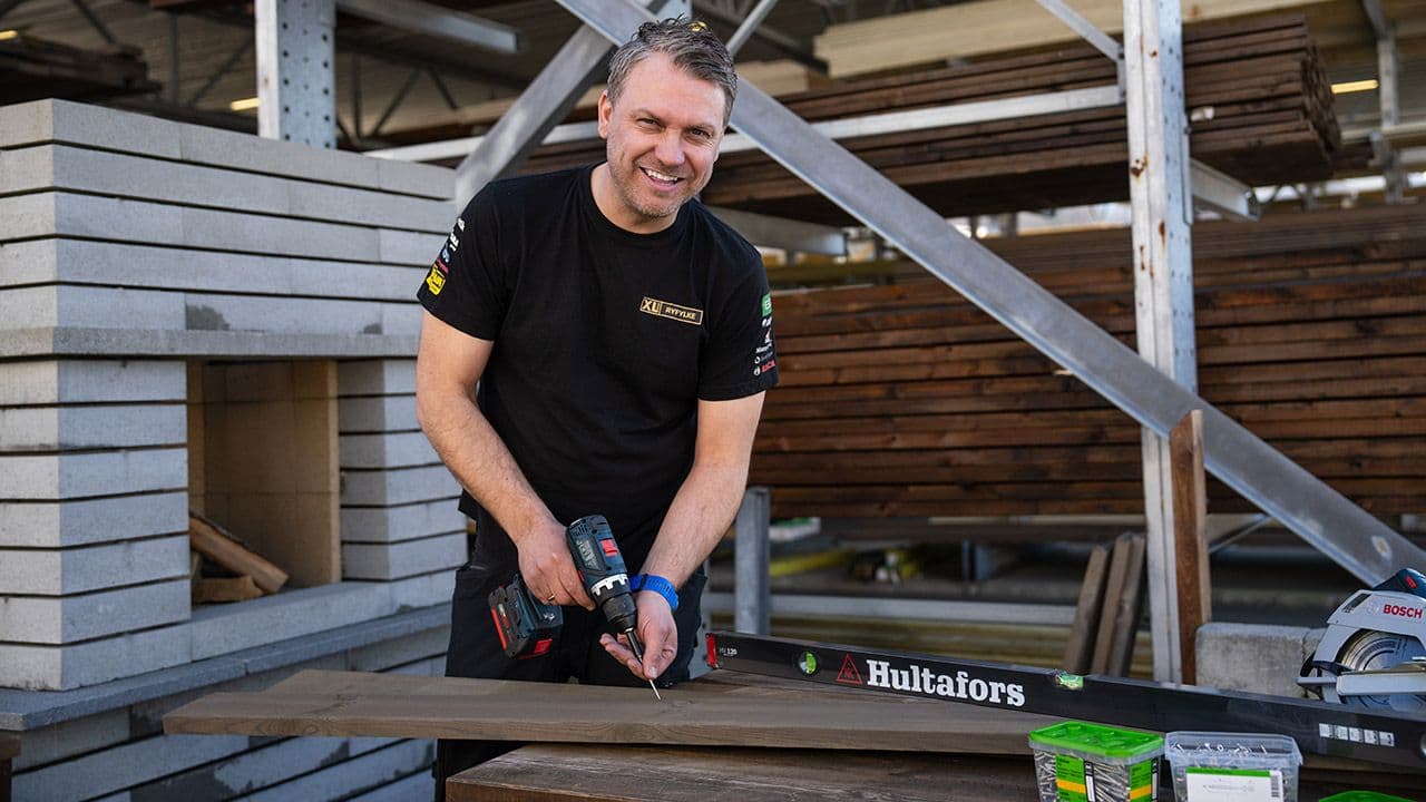 XL-BYGG ansatt smiler mot kamera og viser verktøyene du trenger for å bygge terrasse.