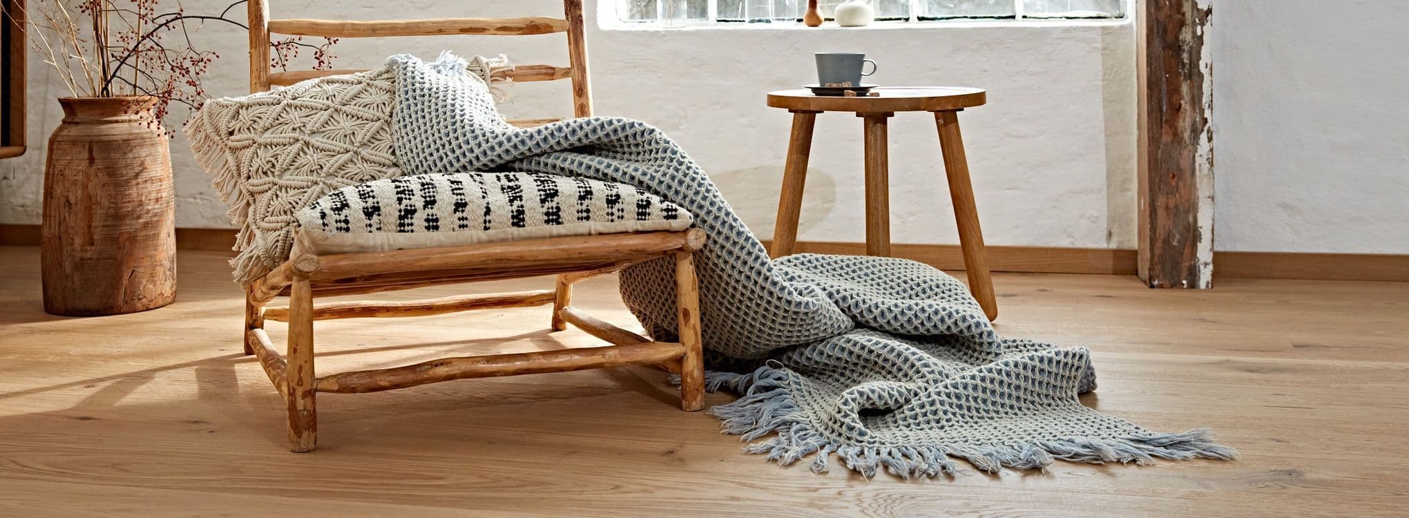 Rustikk trebenk med puter og pledd i naturlige toner på lyst eikegulv.